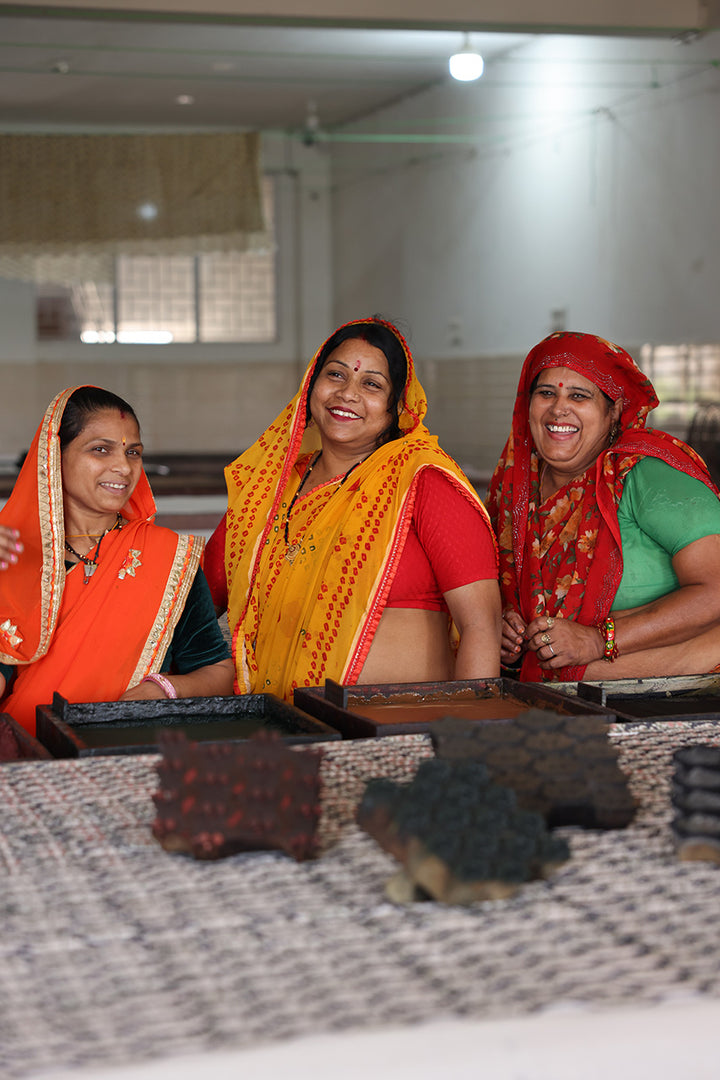 Three smiling Indian women artisans in the Daughters of India workshop who handcraft the Bhoomi Dress fabrics, wearing traditional colourful attire with block printing tools in foreground